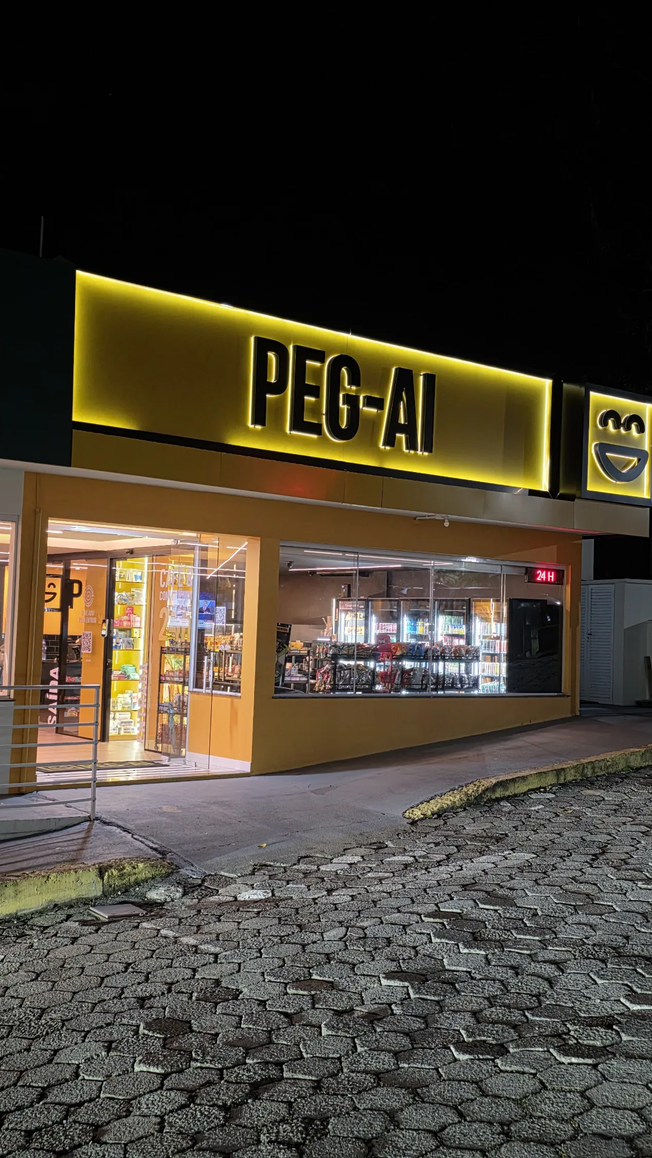Loja PEG-AI iluminada à noite com letreiro grande e vitrine de produtos visíveis, operação 24h