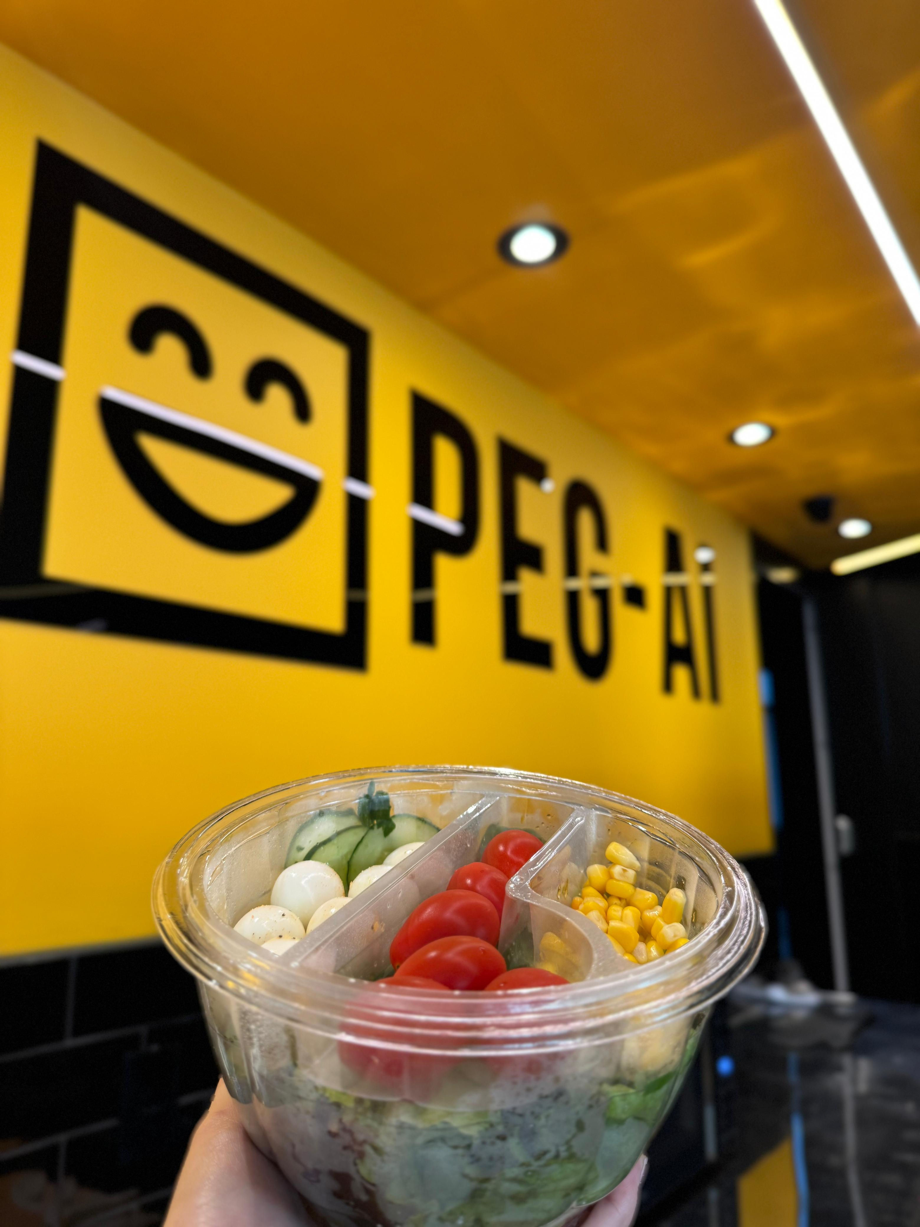 Pote de salada fresca da PEG-AI, com mascote da marca ao fundo, mostrando opção de alimentação saudável na loja