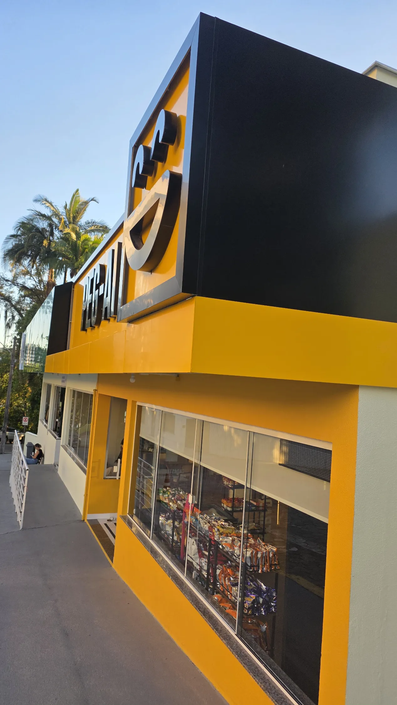 Fachada lateral da loja PEG-AI em perspectiva, com letreiro grande em destaque, palmeiras ao fundo e produtos visíveis pela vitrine